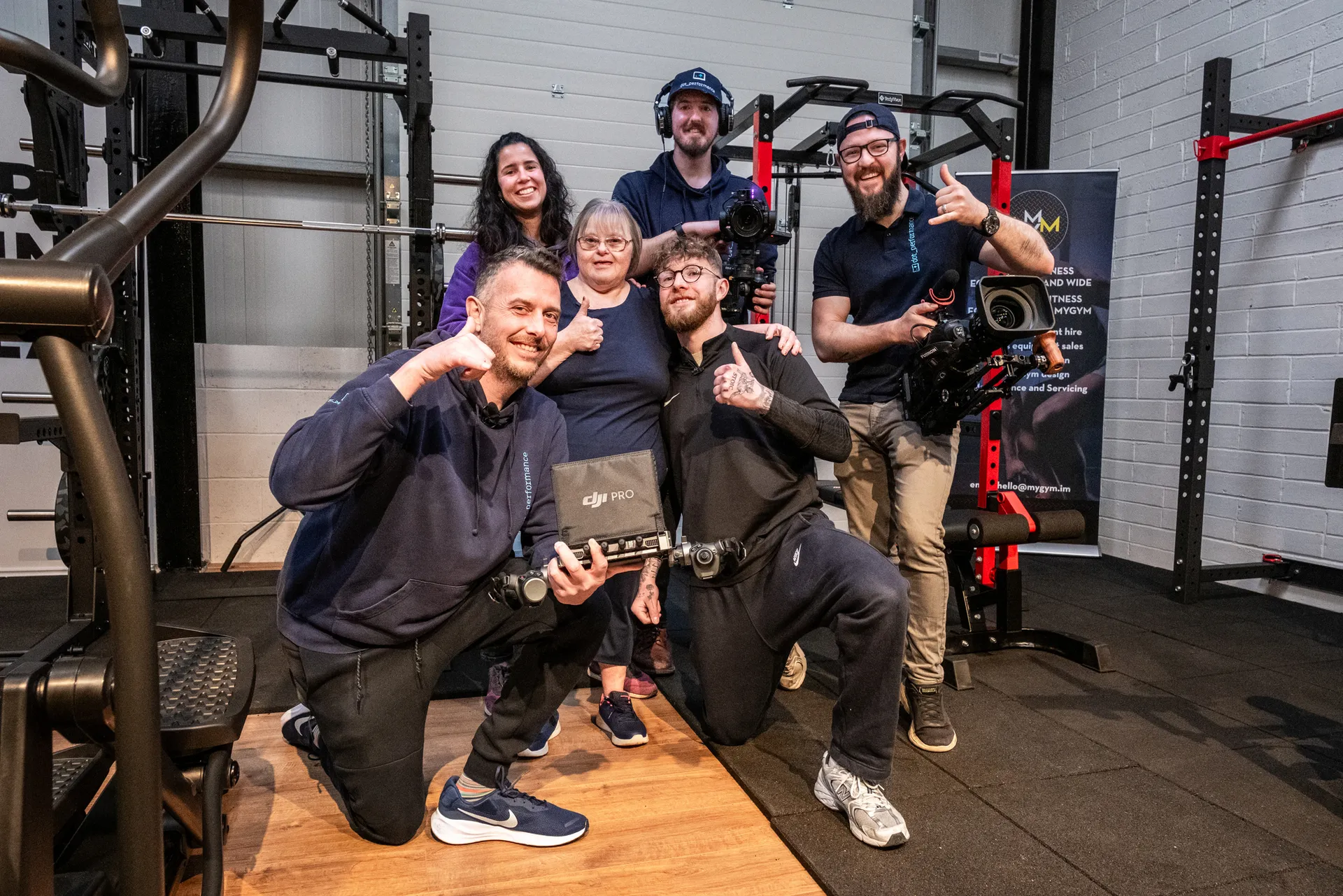 A group of five production crew and gym staff posing with a woman with Down's syndrome in a gym, smiling and giving thumbs up, with DJI Pro camera equipment and a professional video camera visible in a weight training area